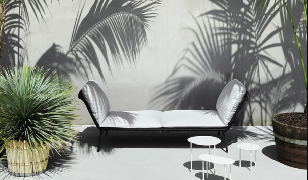 Daydream outdoor daybed with black curved frame and white cushions, flanked by potted plants with palm tree shadows on the wall, accompanied by small white side tables in a modern tropical patio setting.