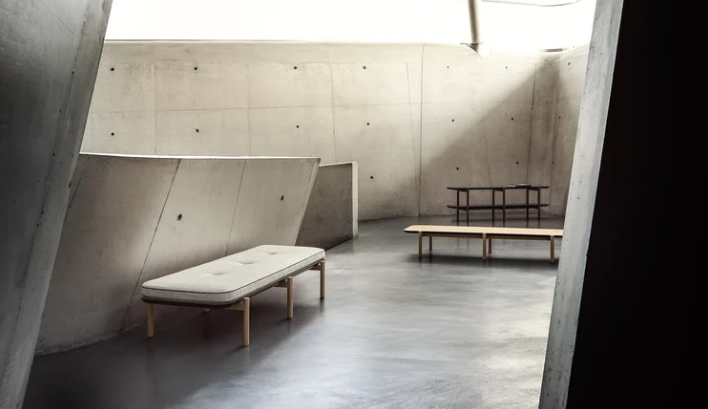 Stepan bench featuring a tufted light-colored fabric cushion supported by a light wood base and slim legs, placed in a spacious, sunlit concrete architectural interior.