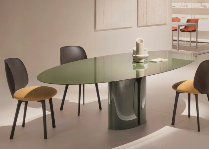 Sleek NVL dining table featuring a glossy, oval green top supported by a central sculptural base, surrounded by upholstered dining chairs in a bright, minimalist showroom.