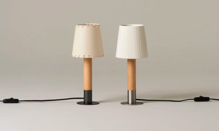 Básica Mínima table lamps featuring cylindrical birch wood stems set on circular metal bases, shown as a pair with stitched leather and pleated fabric shades.