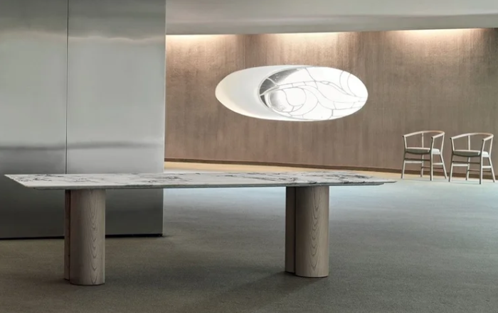 Elly dining table featuring a gray veined marble top supported by cylindrical light wood legs, positioned beneath a sculptural oval pendant light in a minimalist dining room with two white curved-back chairs and neutral-toned walls.