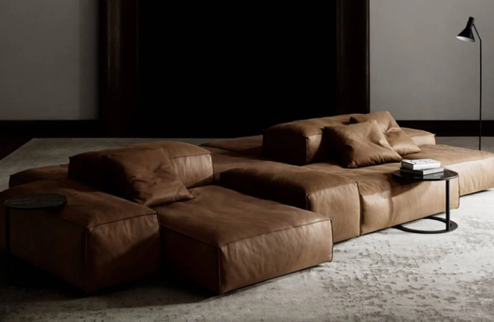 Extrasoft modular sectional sofa upholstered in rich tan leather, configured with low blocky cushions and accent pillows on a textured light rug in a moody interior.