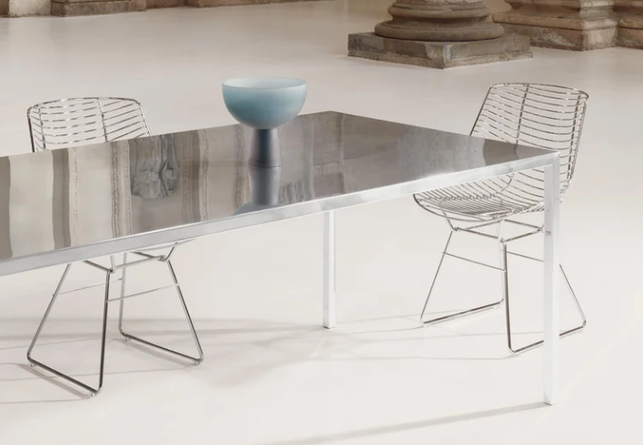 Tense dining table displaying a reflective, polished steel top and matching slender legs, paired with wire dining chairs and a decorative blue bowl in an airy, minimalist space.