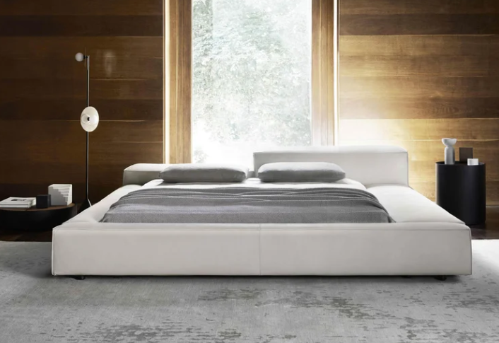 Extrasoft bed featuring a low-profile upholstered white frame with asymmetric headboard cushions, gray linens, and minimalist accent furniture.