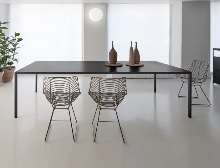 Tense table with a sleek, ultra-thin black tabletop and slim tubular legs, styled with wire dining chairs and ceramic vases in a minimalist, sunlit space.