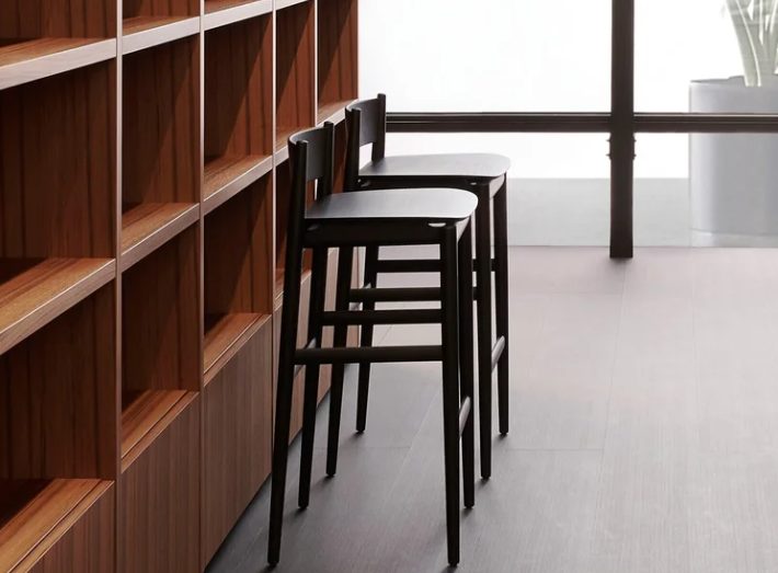 Brina stools featuring dark stained wooden frames with contoured seats and low backrests, lined up beside a rich wood shelving unit in a brightly lit modern interior.