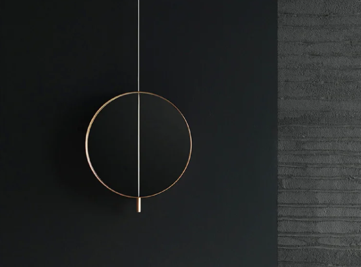 Galileo mirror featuring a circular brass frame bisected by a vertical white rod with a small cylindrical element at the bottom, mounted on a black wall next to textured gray concrete.