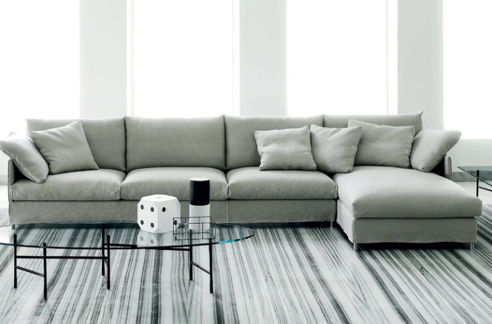 Chemise sofa presenting a spacious light gray sectional layout with plush cushions, styled over a striped marble floor with minimalist glass coffee tables in a bright, sunlit room.
