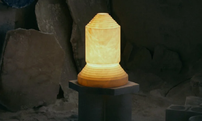 Babel table lamp featuring a sculpted, tiered translucent alabaster vessel glowing softly from within, set on a dark stone pedestal against a raw stone background.