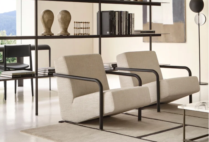 Lullaby lounge chairs featuring beige upholstery with black tubular metal frames and curved armrests, positioned side by side in a modern living space with black metal shelving.