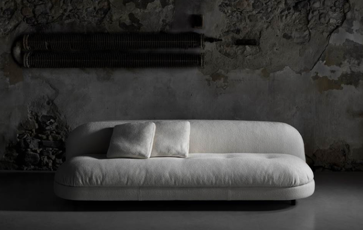 Gala sofa featuring off-white bouclé fabric upholstery with a curved backrest, tufted bench seat, and throw pillows, set against a dark industrial stone wall