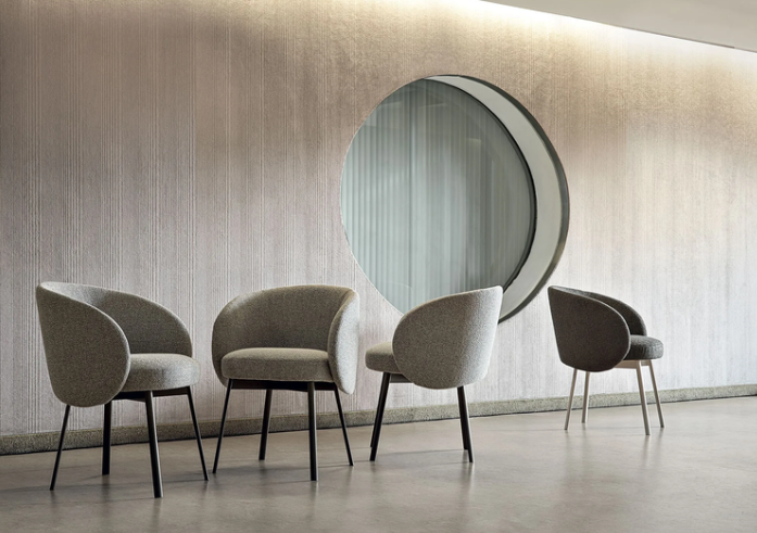 Four Hug dining chairs with curved, enveloping backs upholstered in neutral beige and gray fabrics, arranged in front of a textured wall with a large circular window, featuring thin black metal legs.