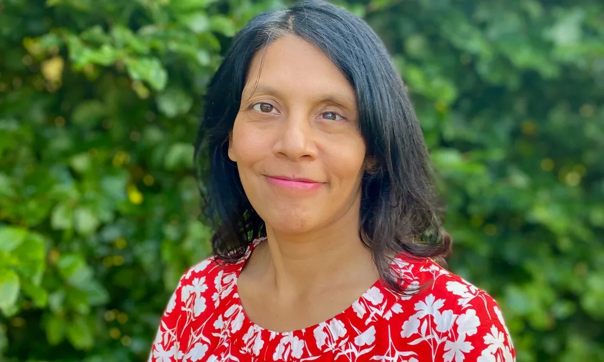 Dr Halima Begum looks at the camera and smiles in this photo taken outside with leaves in background. She wears a red and white top.