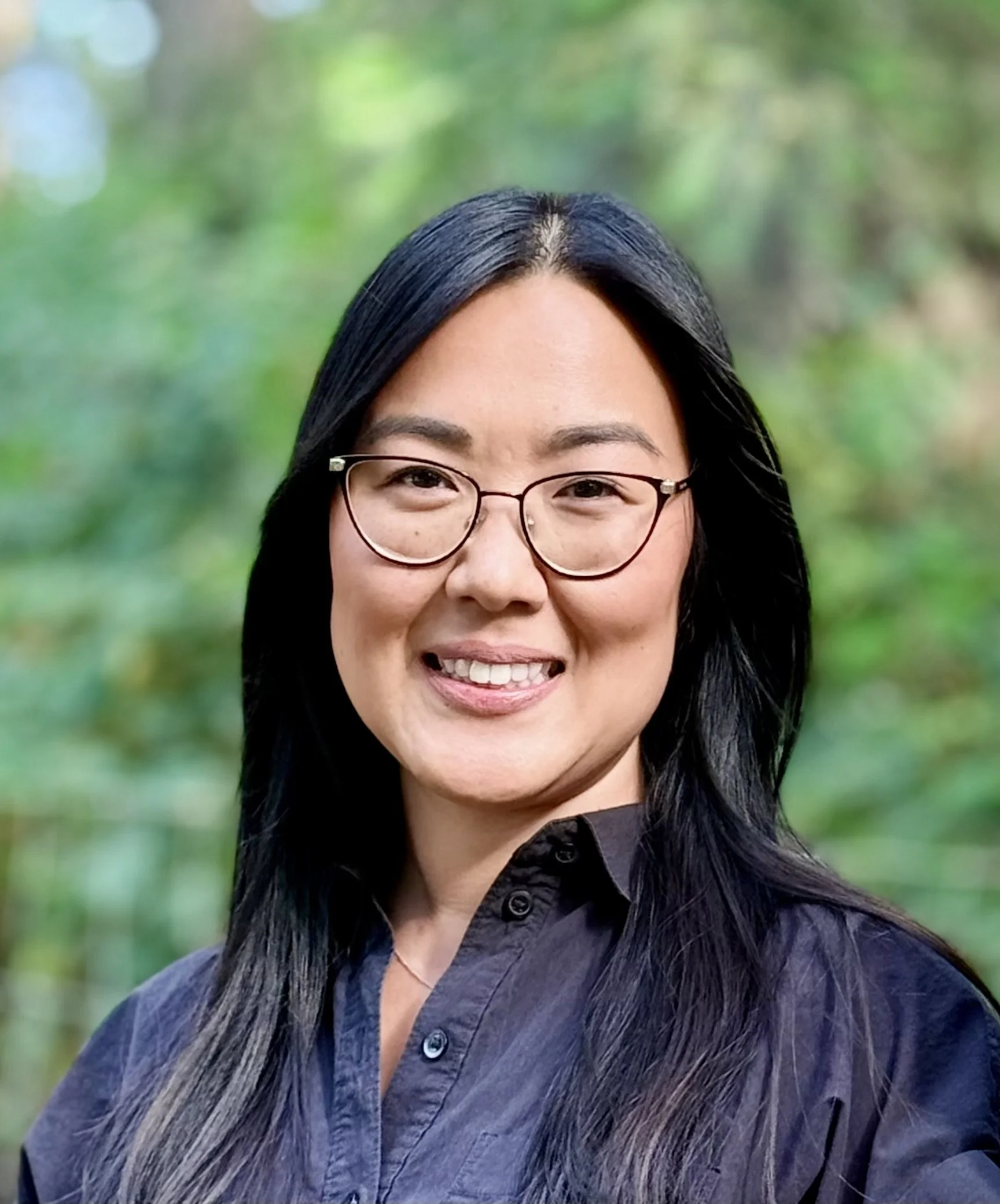 Dr. Eva McCarthy smiling, wearing glasses and a dark shirt, with long dark hair in an outdoor wooded setting.