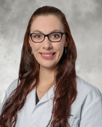 Dr. Atalie Rutherford smiling, wearing black glasses and a white medical coat, with long reddish-brown hair against a gray background.