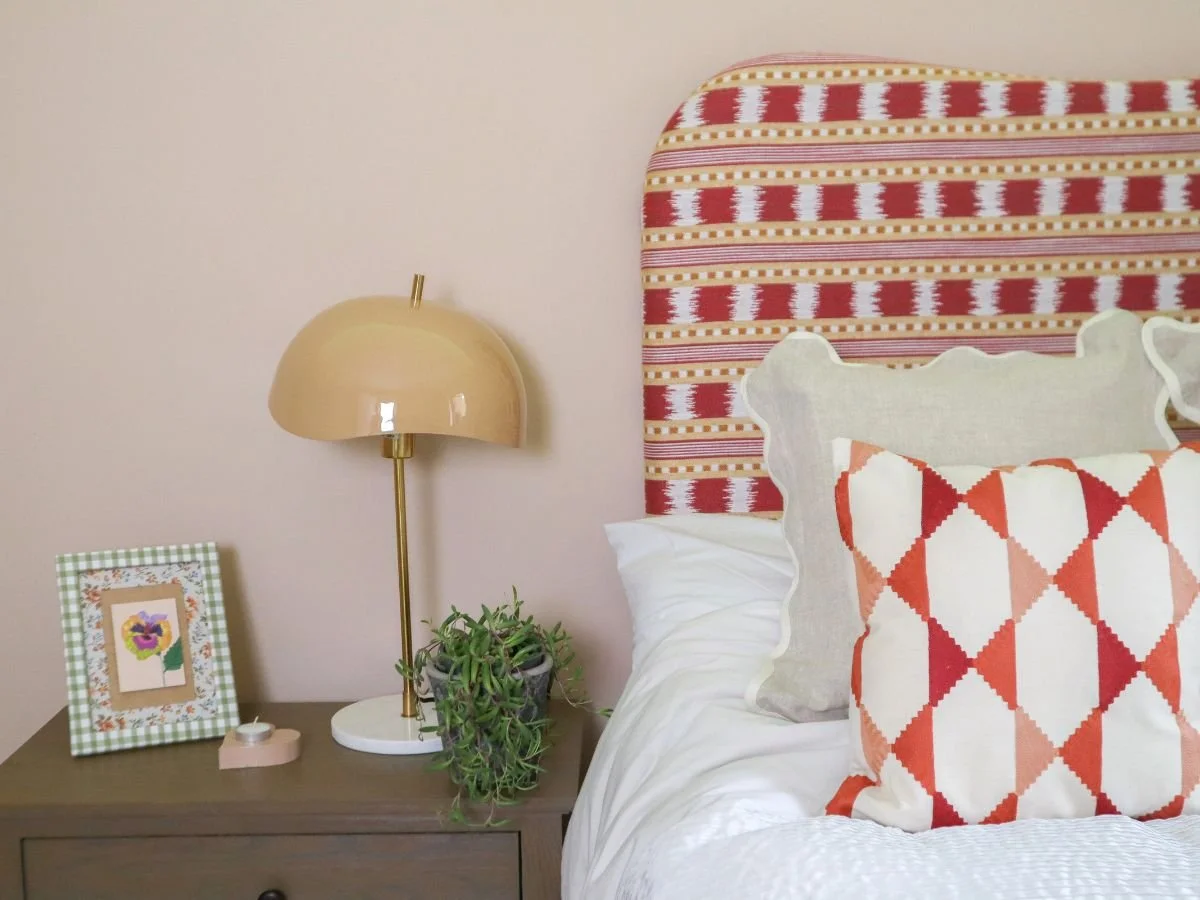 Close-up of a bedroom nightstand with a framed flower picture, beige table lamp, and a small potted plant. Part of a bed with patterned pillows and upholstered headboard are visible.