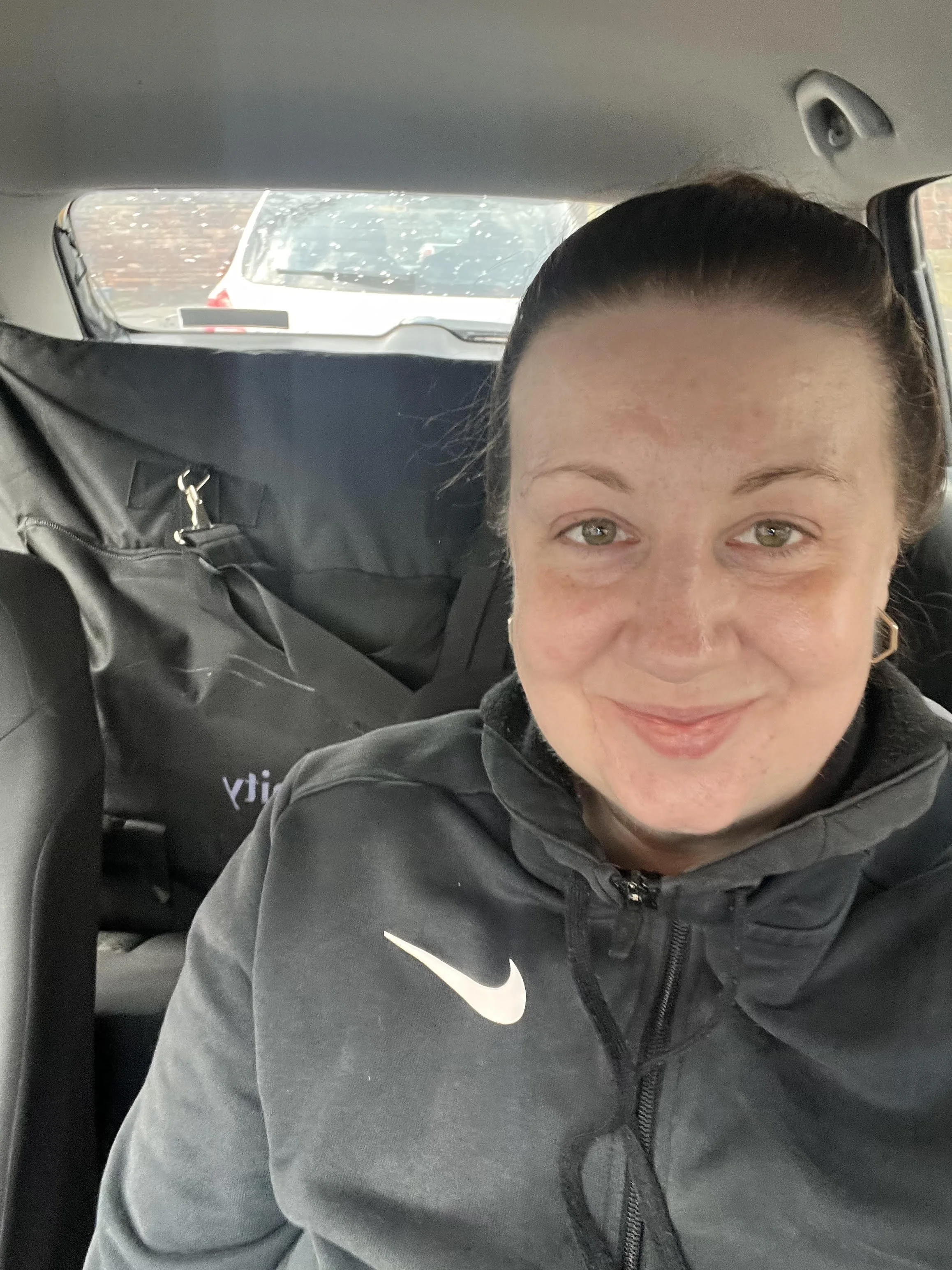 A smiling woman with dark hair tied back, wearing a dark Nike jacket, sitting in the backseat of a car with a black bag behind her and other cars visible through the rear window.