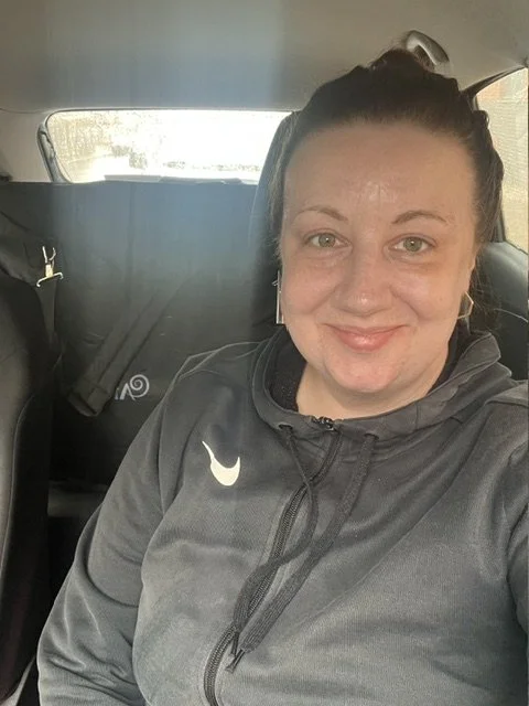 A woman with dark hair tied back, wearing a dark gray Nike zip-up jacket, sitting in a car and smiling at the camera.