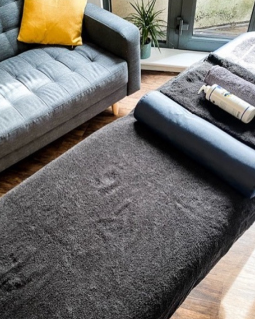 Close-up of a massage or therapy table covered with a dark grey towel, with a rolled-up black towel and a small bottle on top, in a room with a grey sofa, yellow pillow, and a window showing a garden outside.