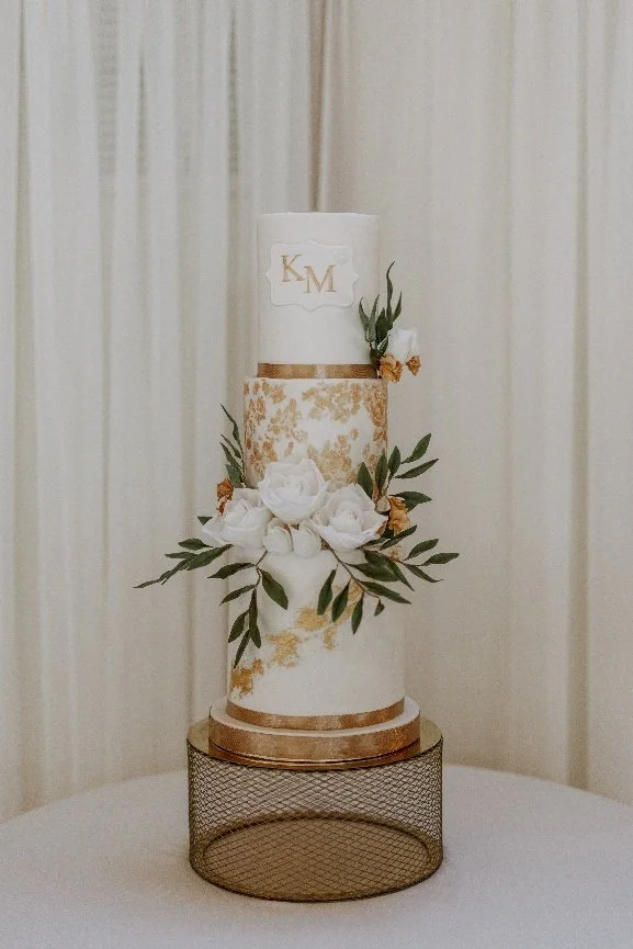 Four-tier white and gold wedding cake with hand-made sugar roses and a gold monogram, made for a wedding at Old Kent Barn