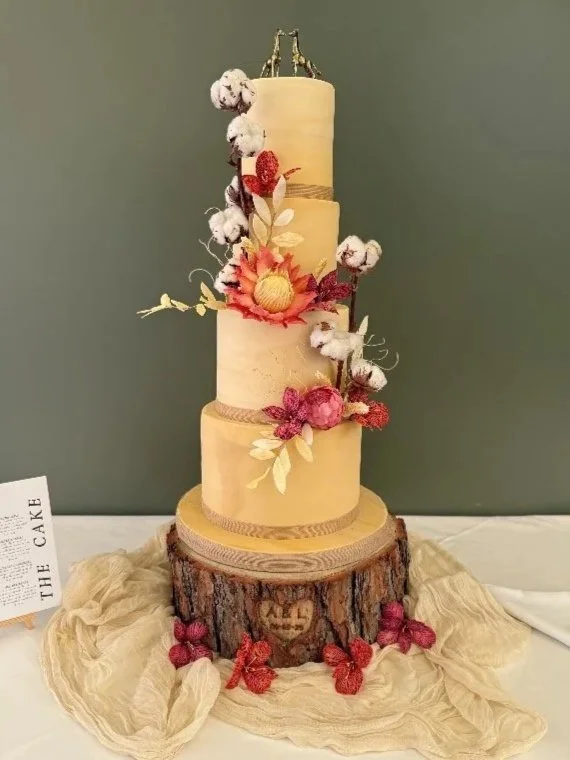 Four-tier marbled wedding cake with sugar proteas and cotton flowers, topped with gold giraffes, on a wooden slice stand, made for a wedding at Port Lympne