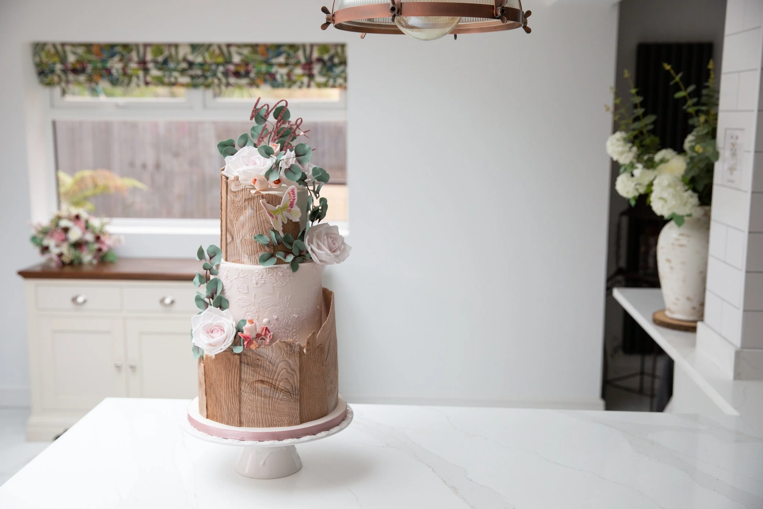Three-tier cake with floral and eucalyptus decorations on a kitchen counter.