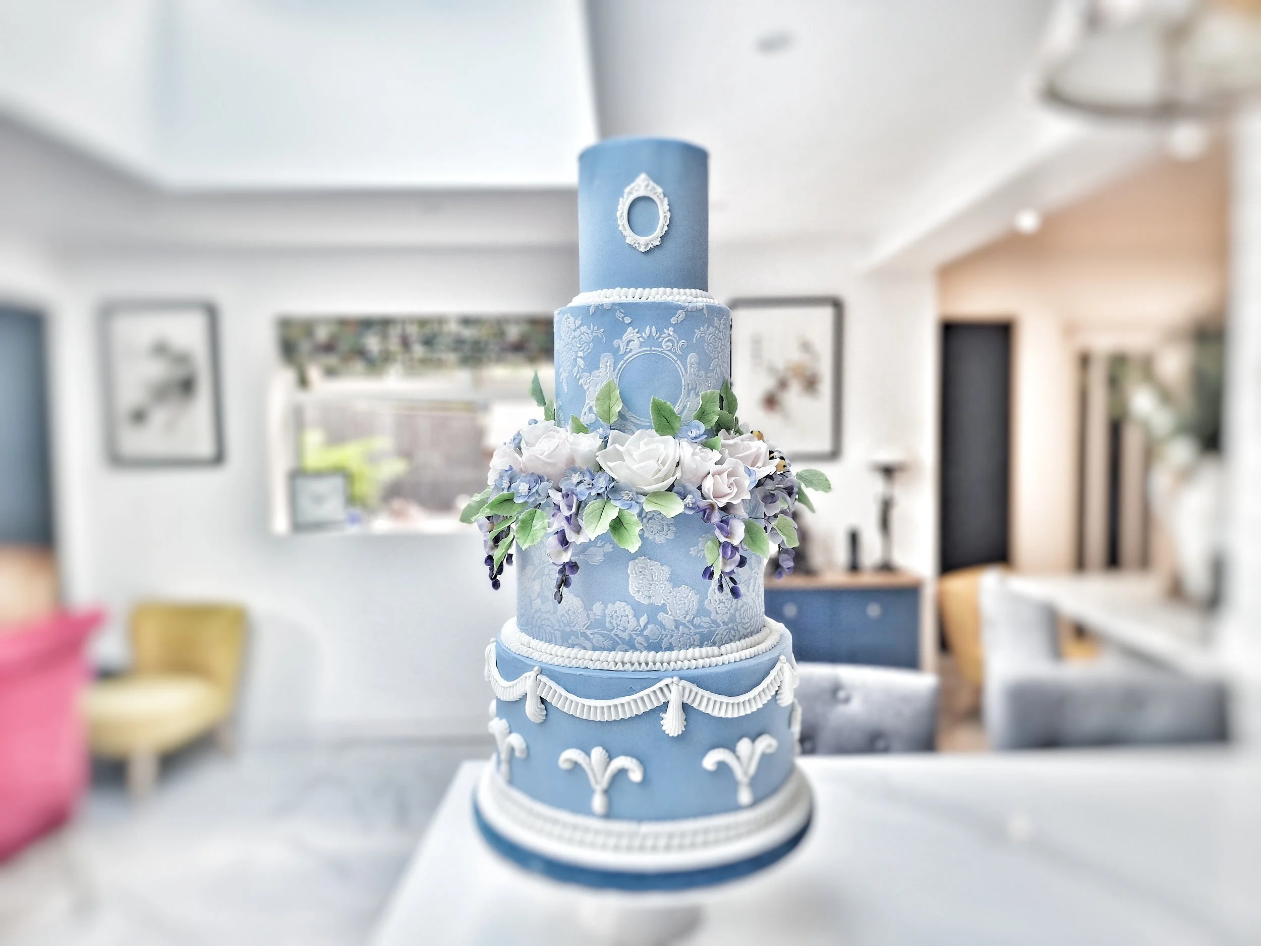 A tall, multi-tiered blue wedding cake with white decorative piping, floral accents, and a flower-shaped cake topper, displayed in a softly lit room.