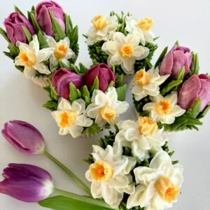 Cupcakes decorated with flowers resembling tulips and daffodils, with real purple tulips nearby on a white background.