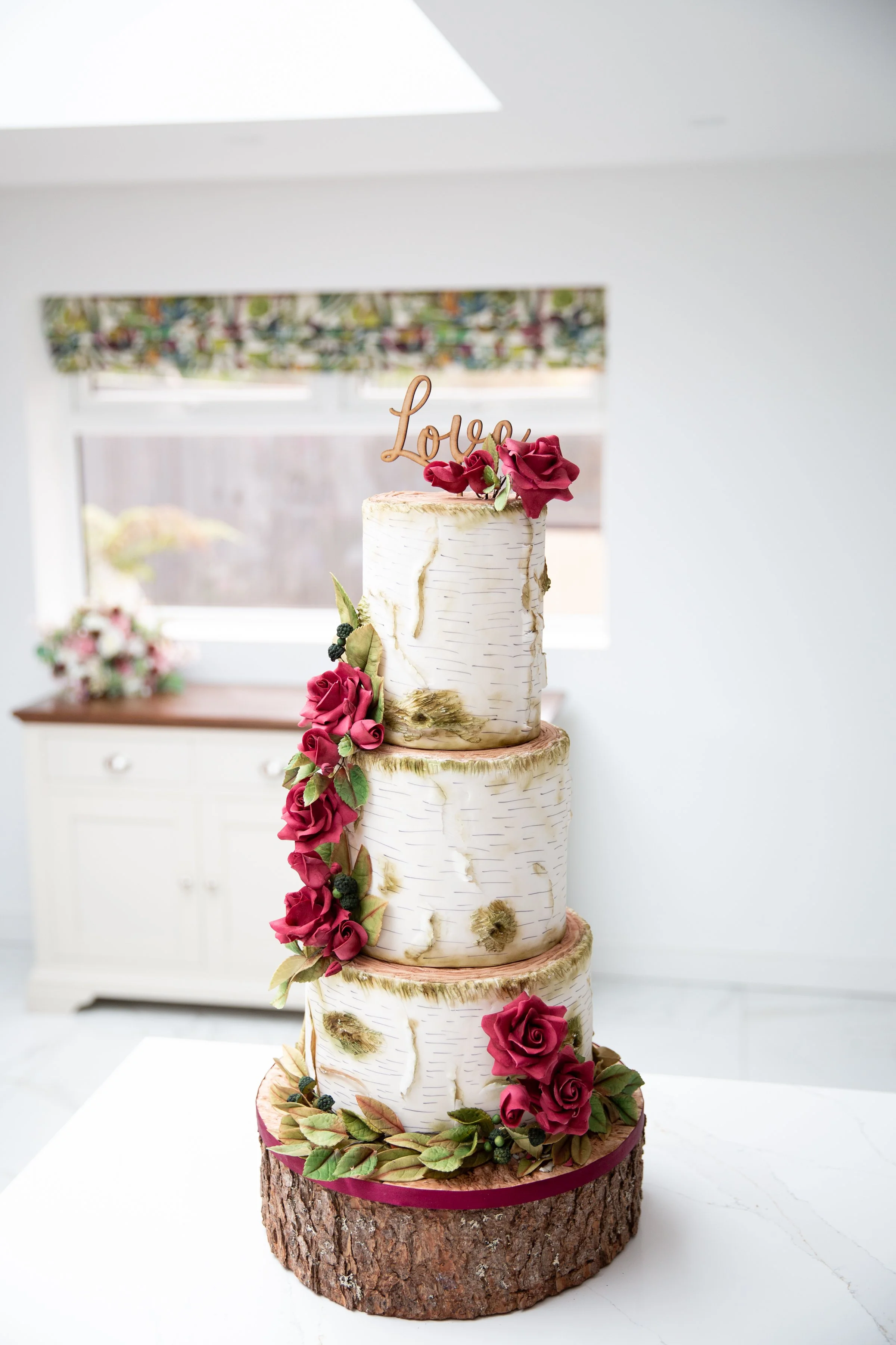 Three-tier birch-themed wedding cake with red roses and "Love" topper, displayed in a bright room.