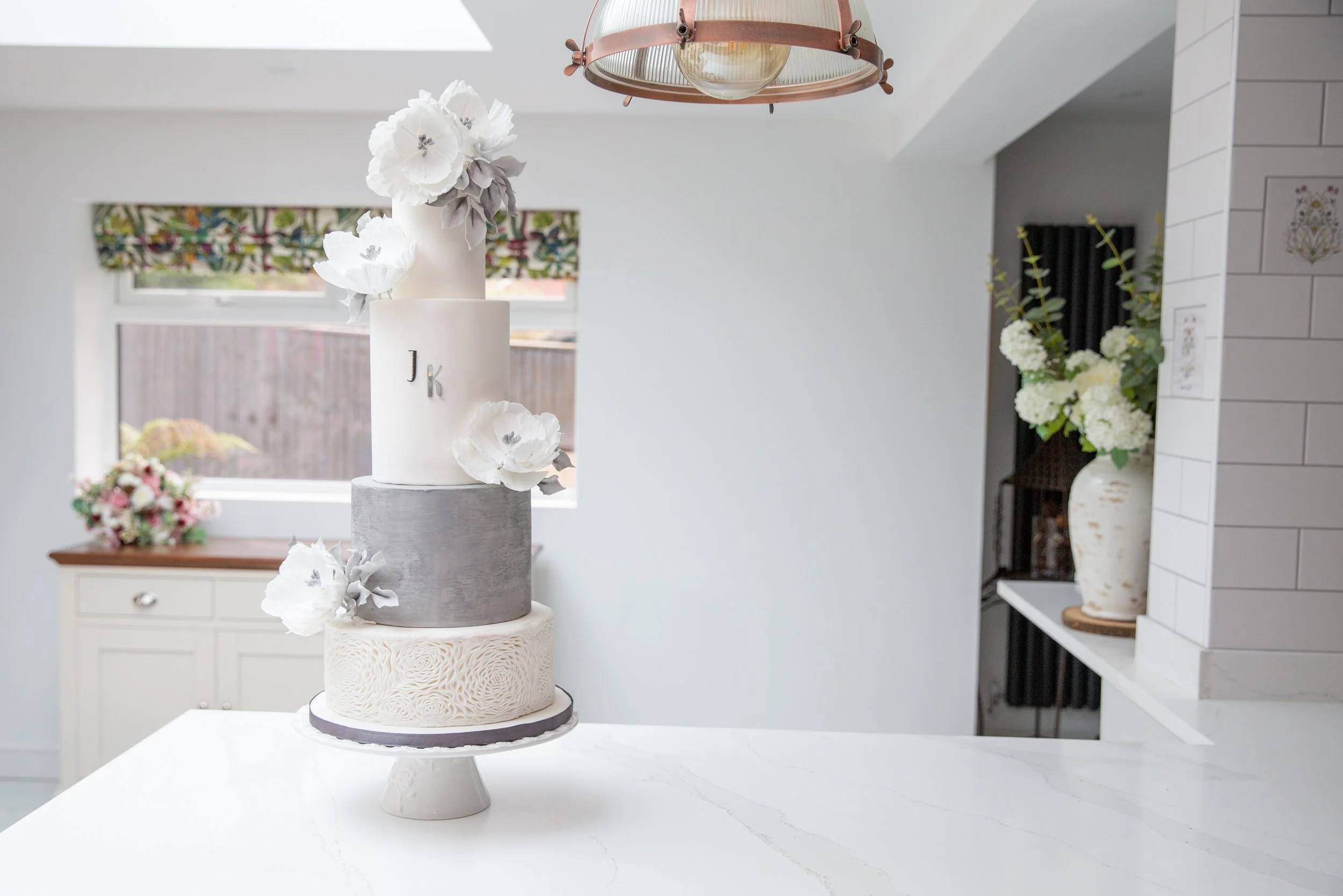 Three-tiered wedding cake with white and gray fondant, decorated with white flowers, on a stand in a modern kitchen.