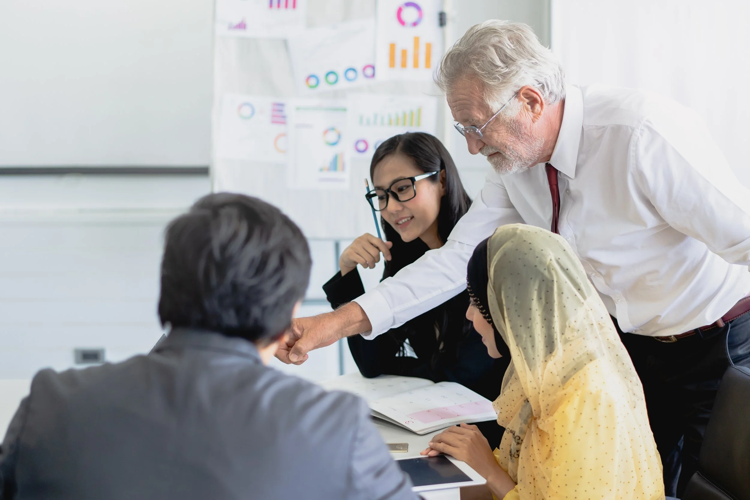 A group of people having a business meeting, with one person pointing at something on a table, and charts on the wall behind them.