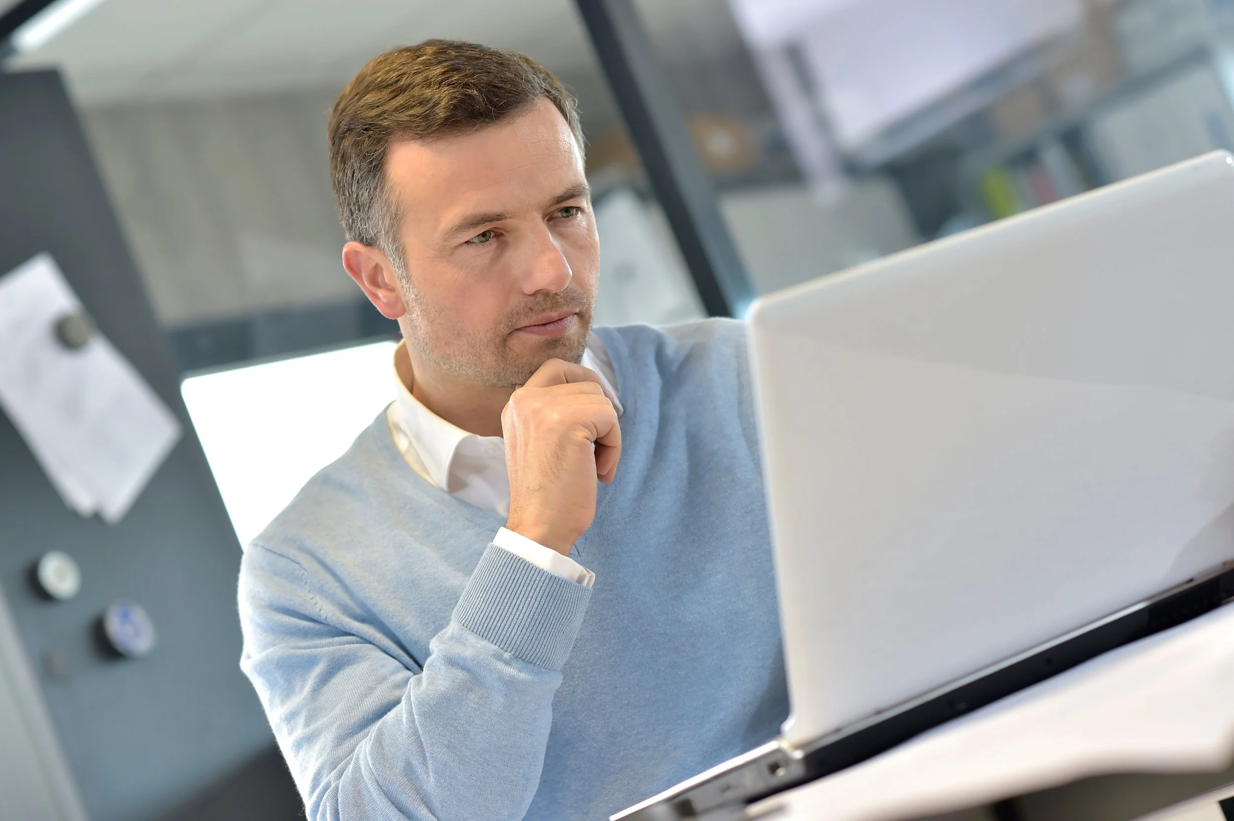Man in a light blue sweater thinking while looking at a laptop in an office setting.
