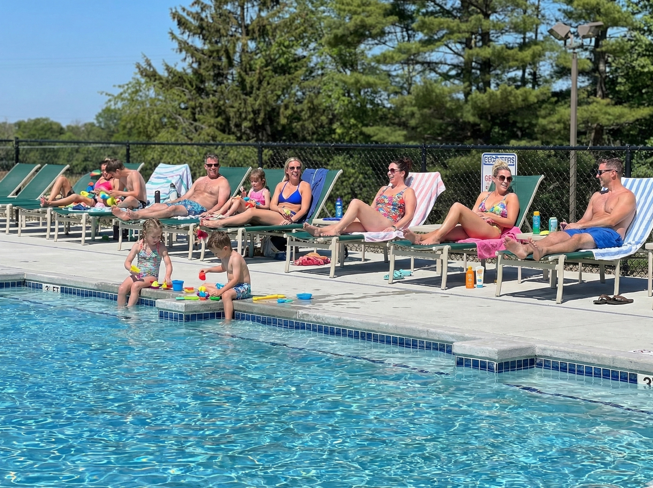 View of a pool from the side with a concrete deck and many green lawn chairs surrounding it.