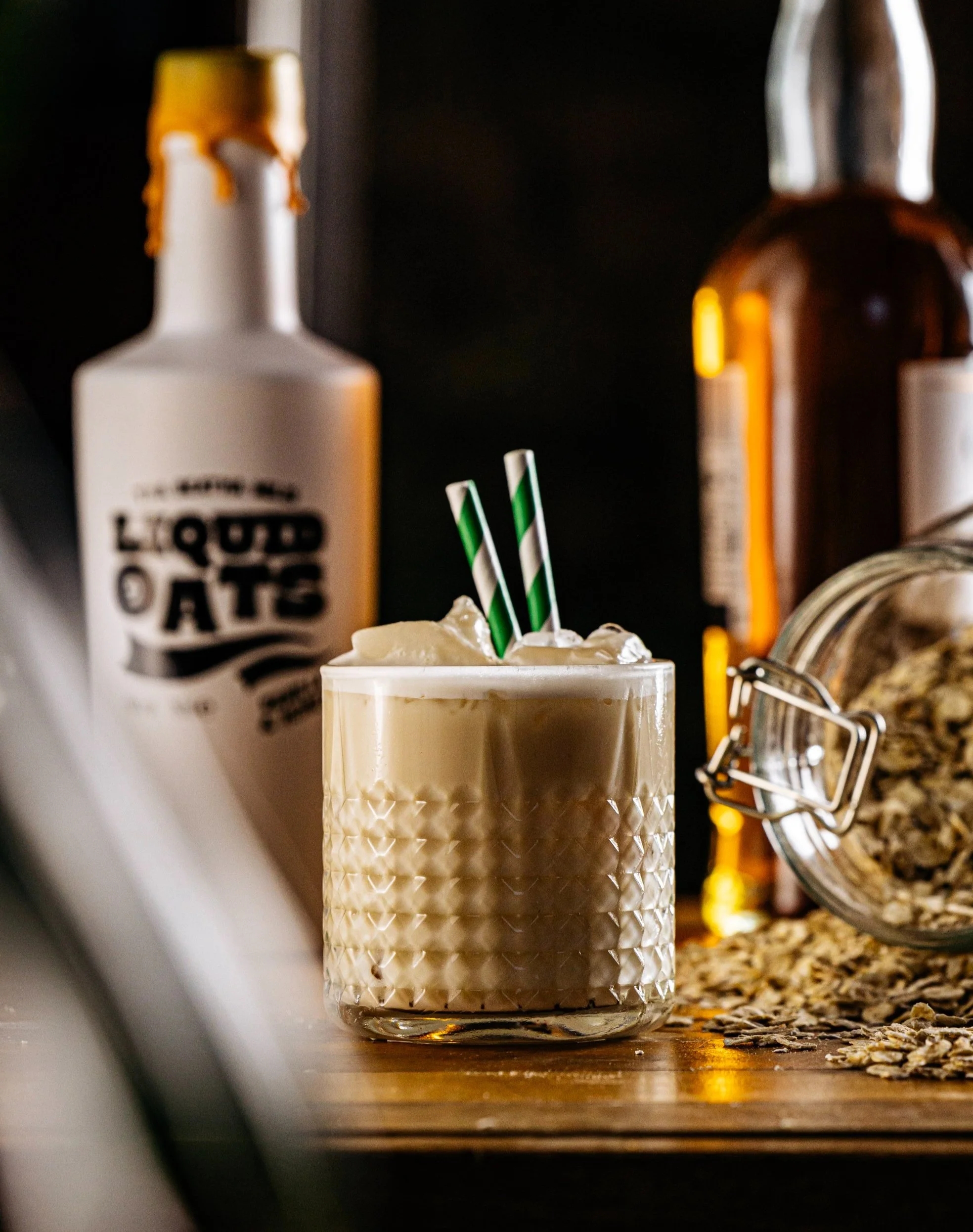 A glass of creamy beverage topped with whipped cream and two green and white striped straws on a wooden surface, with bottles and a jar of oats in the background.