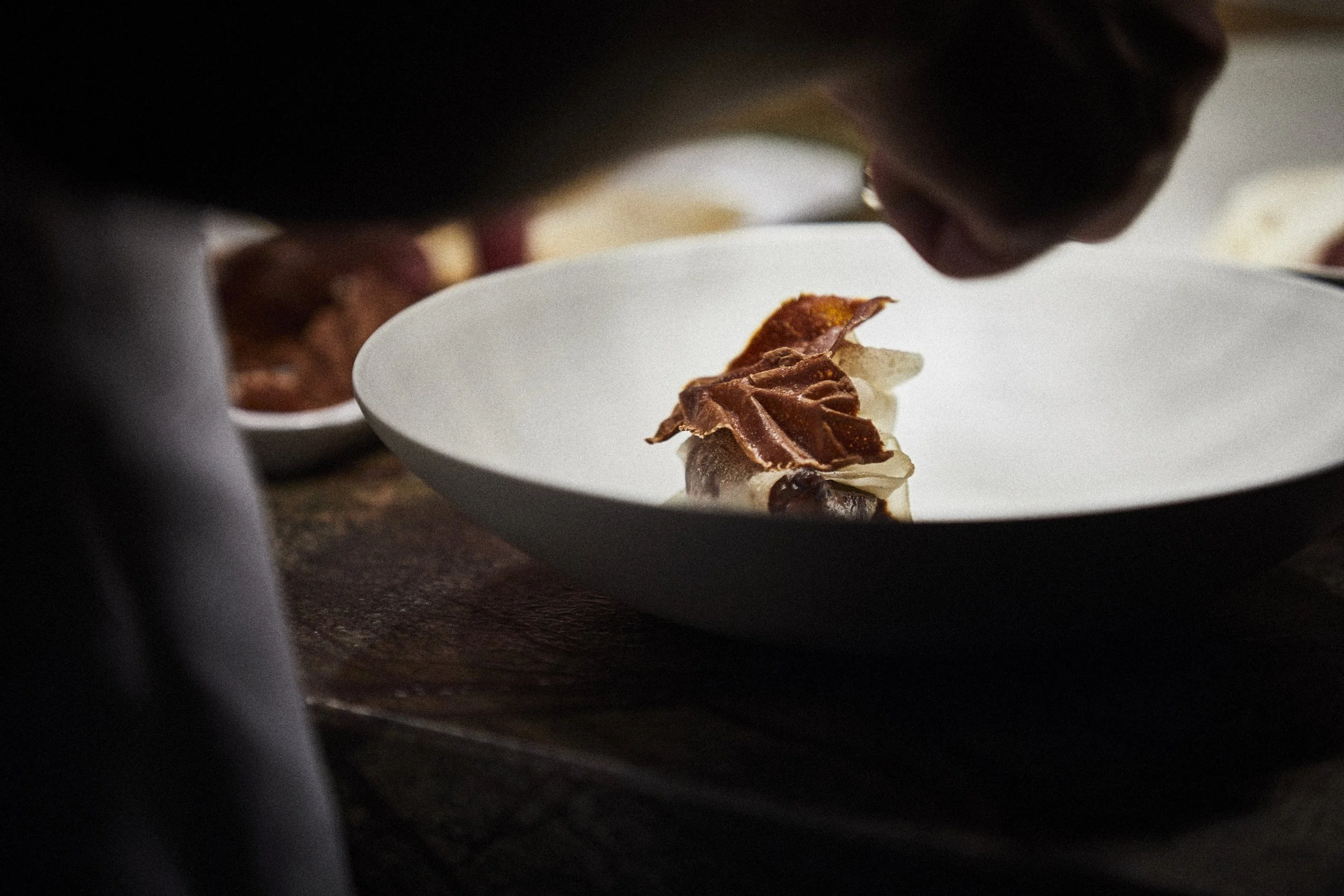 Nahaufnahme eines Küchenchefs, der ein Gericht auf einem weißen Teller anrichtet, mit mehreren Tellern im Hintergrund.
