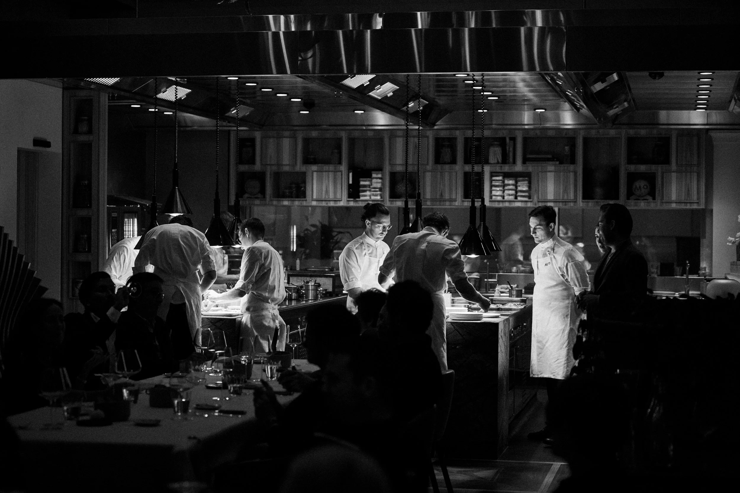Schwarzes und weißes Bild eines Restaurantkochens bei der Zubereitung von Speisen in einer offenen Küche, mit Gästen, die am Tisch sitzen.