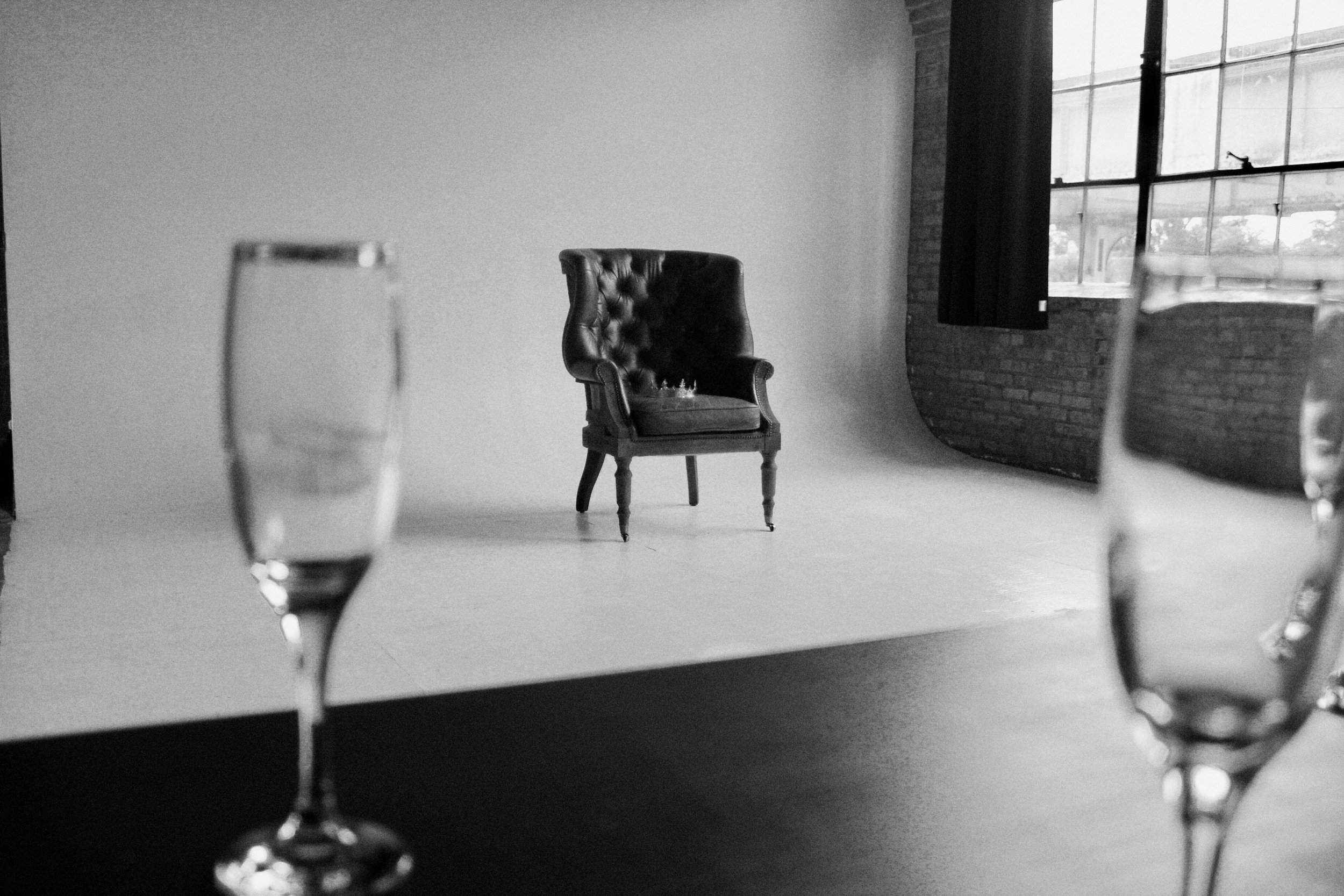 Black and white photo of two wine glasses in the foreground viewed from behind, with a vintage leather armchair and a chess set on it in the background near a brick wall and large window.