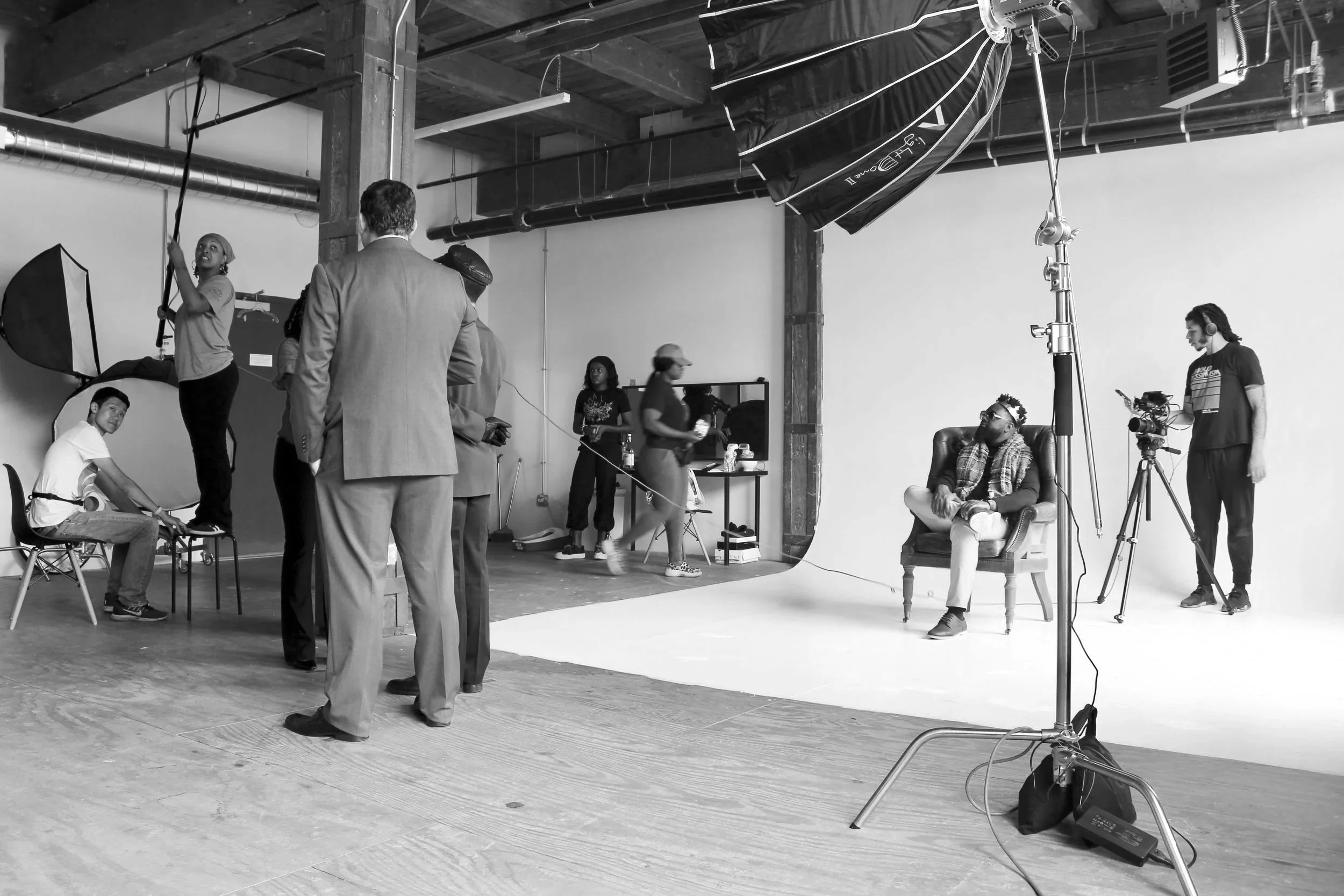 Behind the scenes of a video or photo shoot in a studio. Crew members setting up equipment and a man sitting in an armchair in front of a white backdrop.