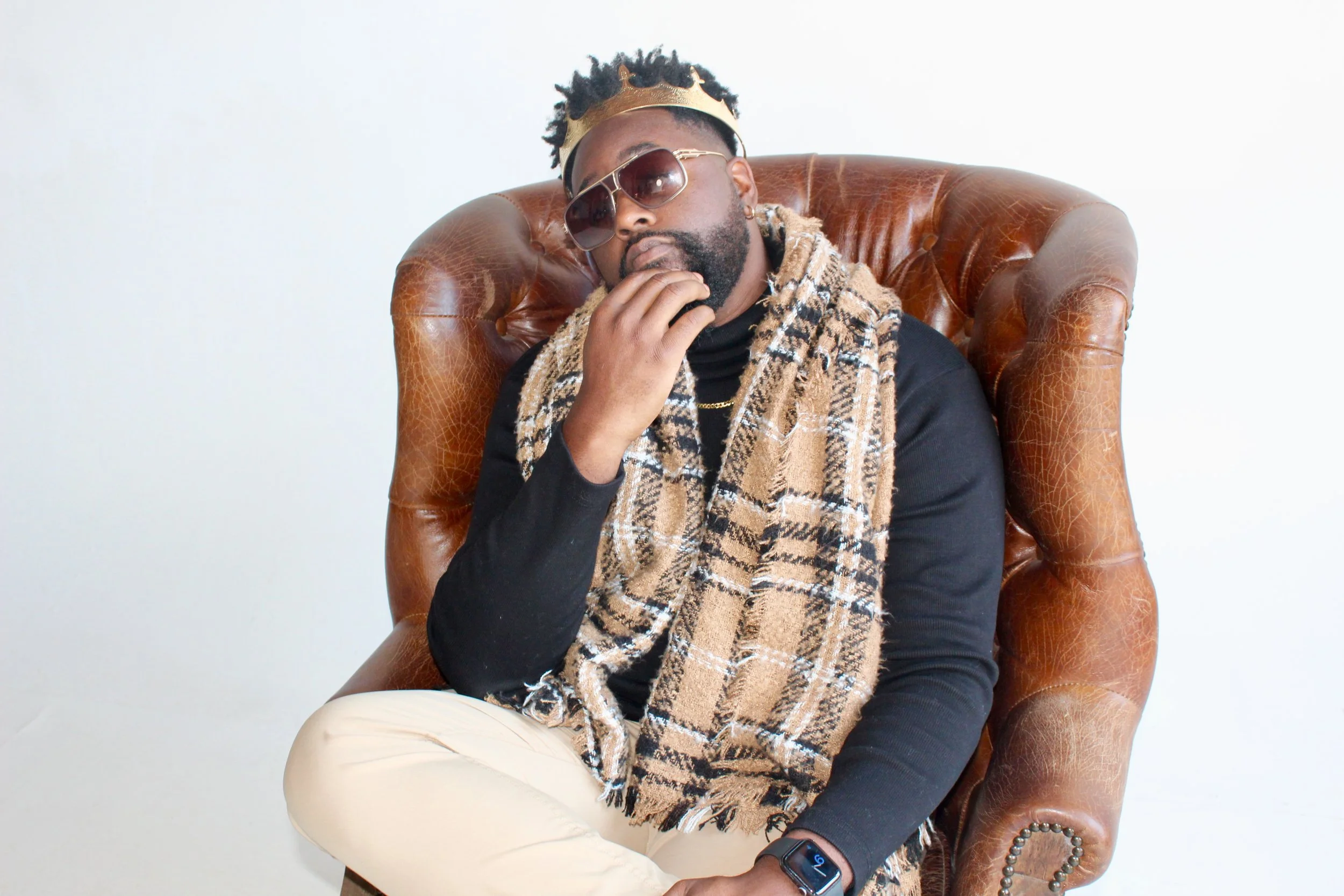 A man wearing sunglasses, a crown headband, and a beige checkered scarf sitting on a brown leather armchair.