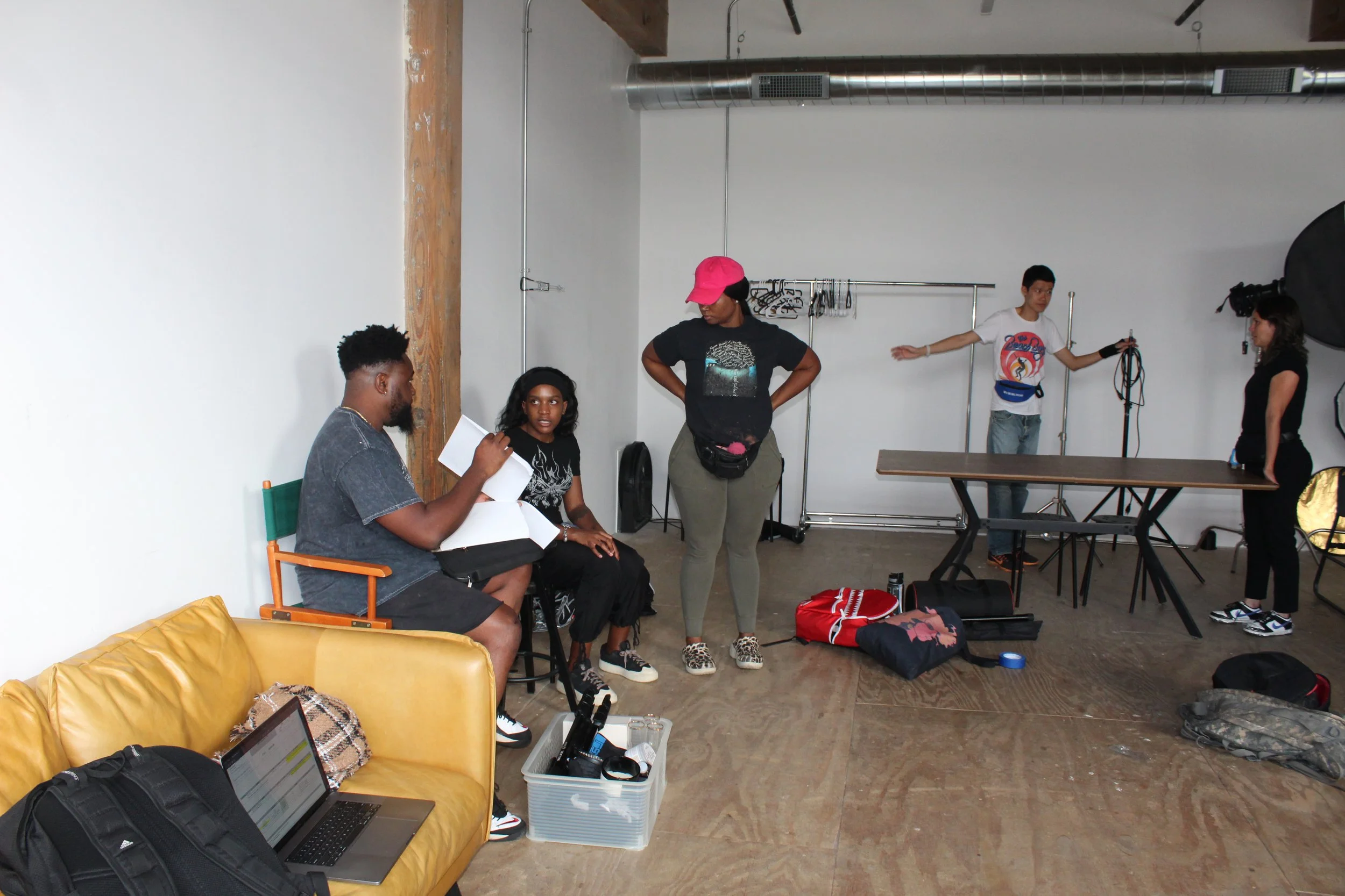 A group of five young people in a photo studio, with some seated on a yellow couch and others standing. The studio has a plain wall, wooden floor, and photography equipment.