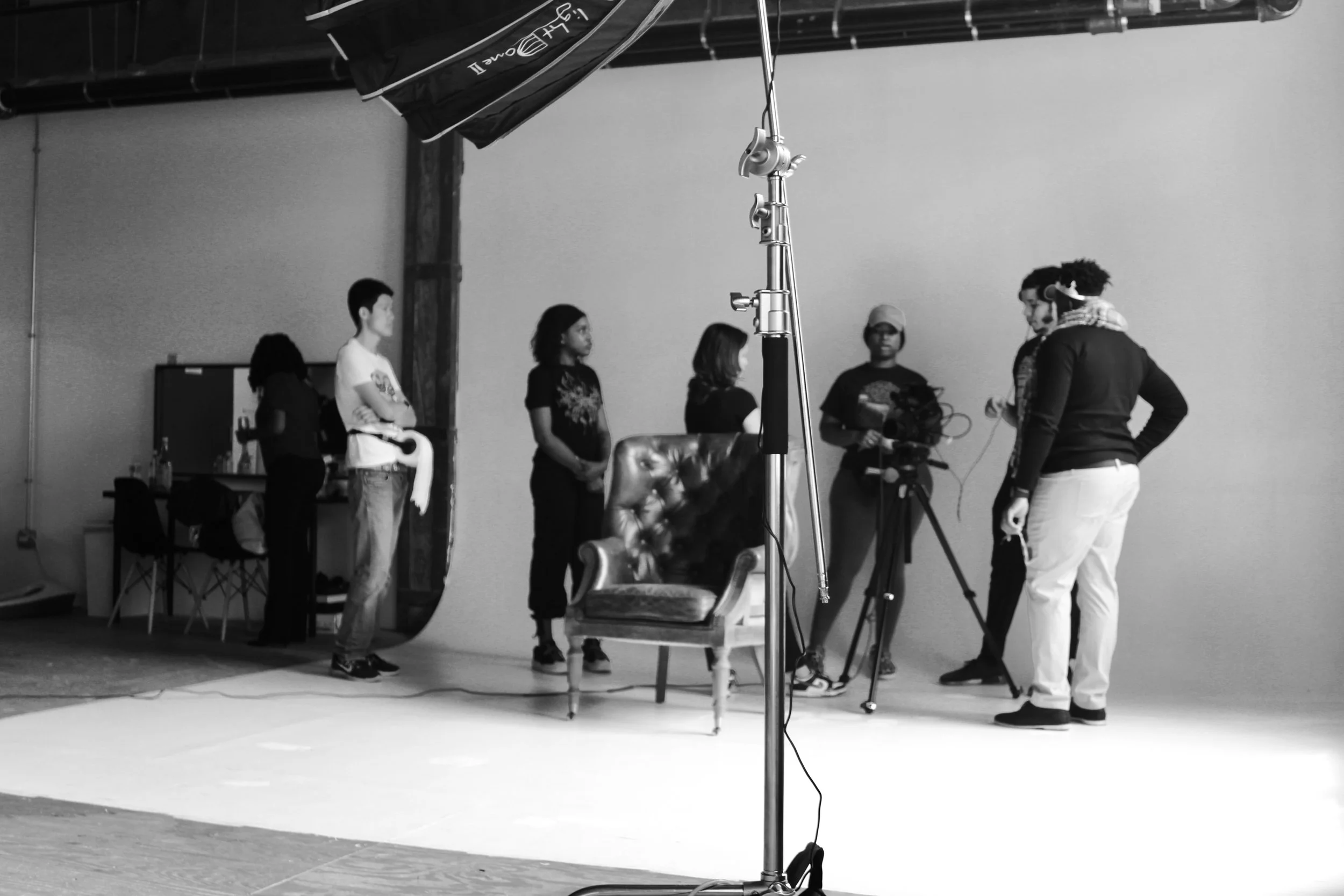 Black and white photo of a behind-the-scenes view of a photo or video shoot. Several people are gathered around a camera and a vintage-style armchair, with some standing and others preparing equipment. A large umbrella reflector is visible overhead, 