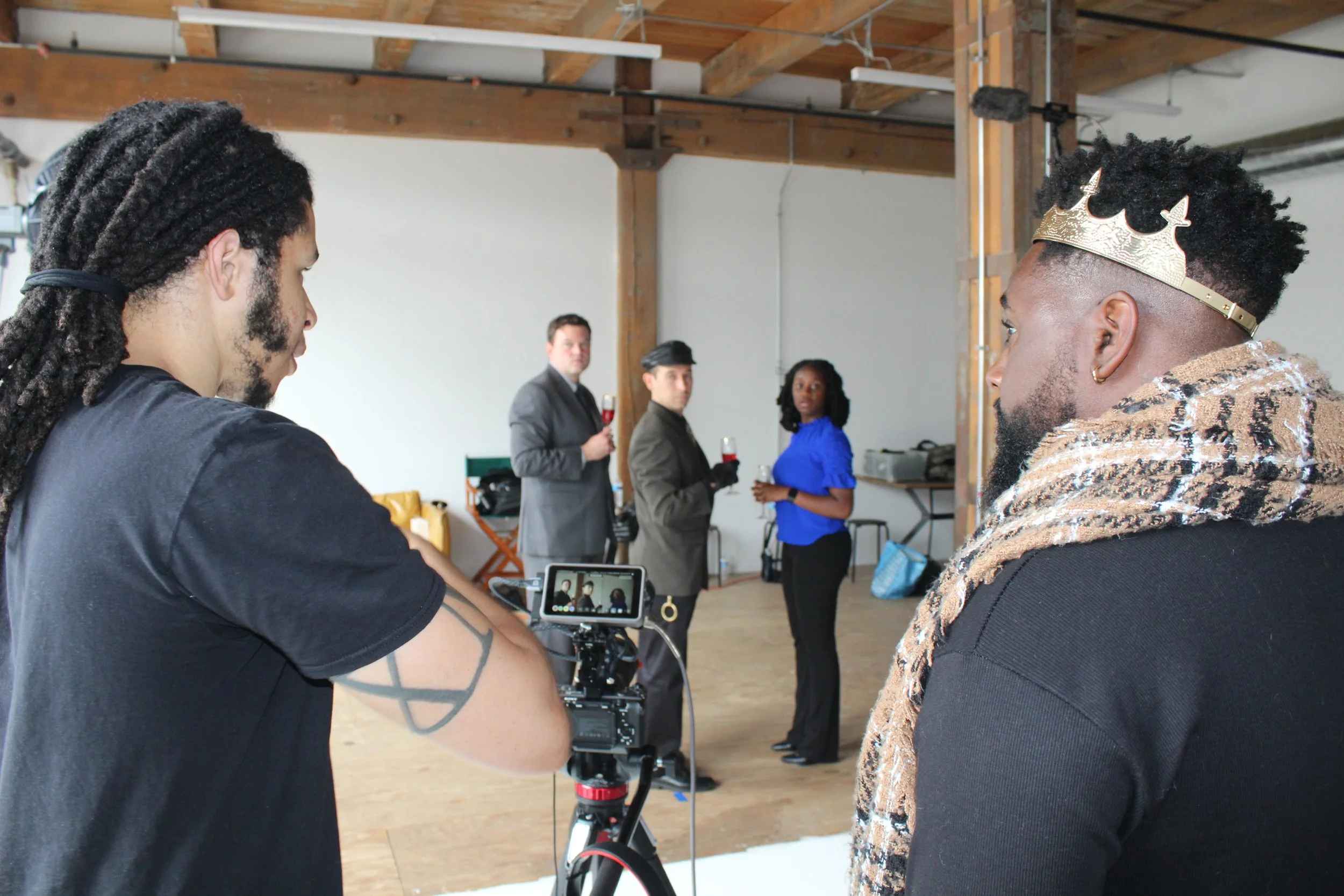 Film crew filming a scene with four actors in a studio with wooden beams. One actor wears a crown and a leopard-print scarf; others hold glasses of red wine.