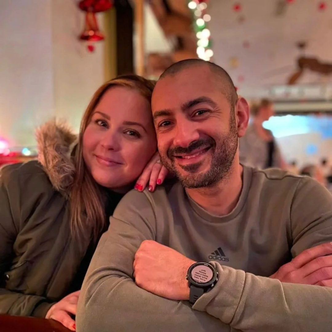 A man and woman smiling, sitting closely at a restaurant table. The woman has light skin and long brown hair with a fur-lined hood, and the man has a shaved head, beard, light skin, and is wearing a beige Adidas shirt and a smartwatch. The background has blurred colorful lights and decorations, indicating a festive holiday atmosphere.