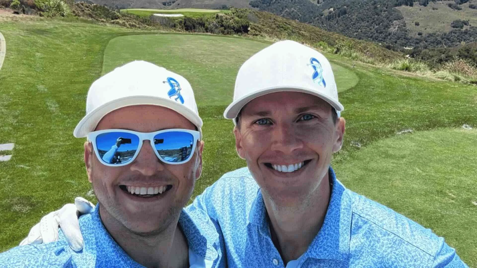 Chris and Markham Rollins smiling on a golf course, wearing white caps and blue patterned shirts, with green hills and a sand trap in the background.