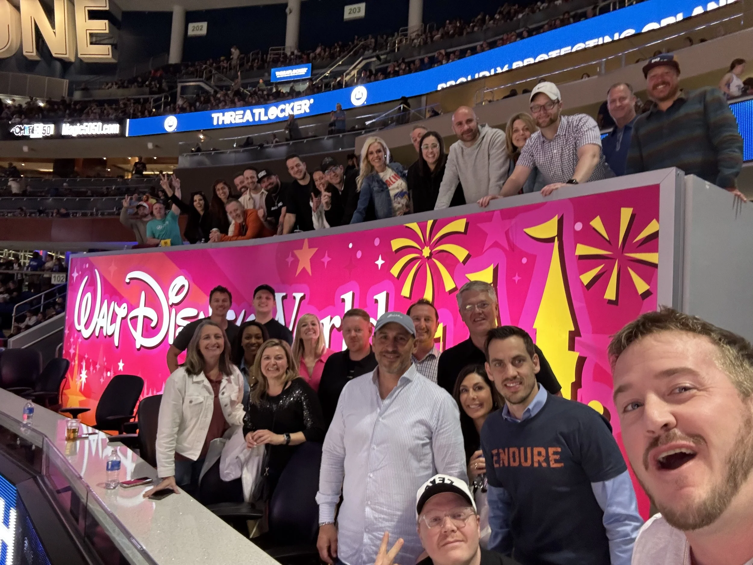 A group of people at a Disney World event, posing for a photo in front of a pink sign with fireworks and castle graphics. The background shows a stadium with more attendees, and a digital billboard displaying advertisements.