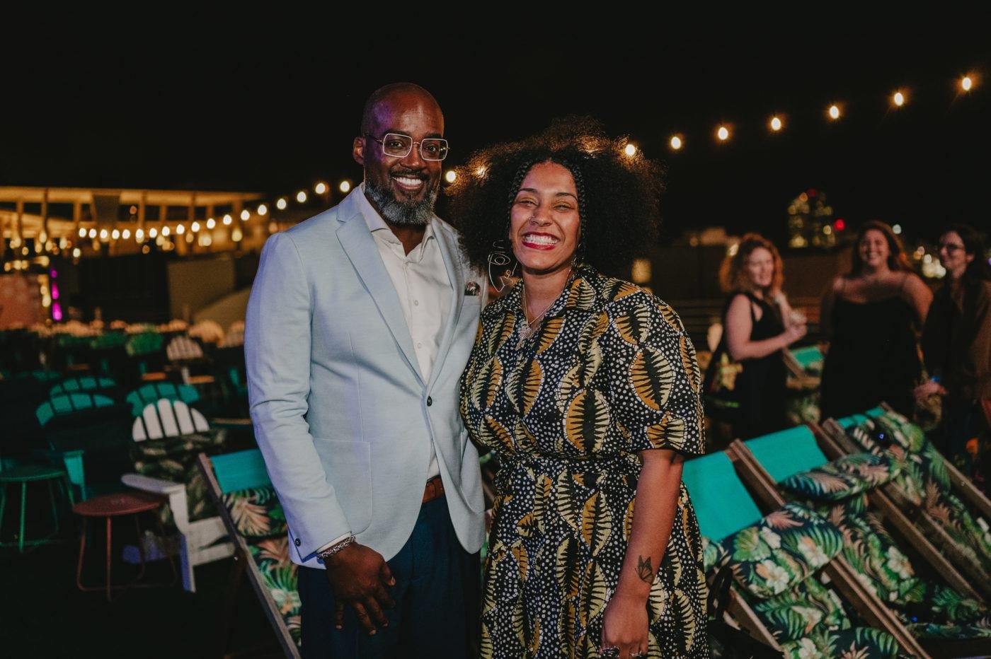 A man and woman smiling at a nighttime outdoor event with string lights, surrounded by people and teal-colored chairs.