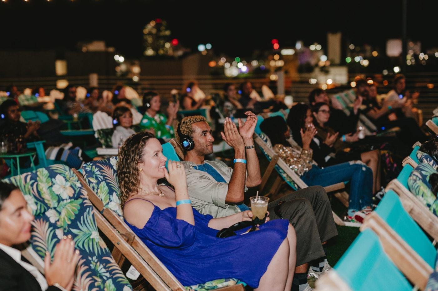 People sitting in deck chairs at a nighttime outdoor event, some wearing headphones and clapping.