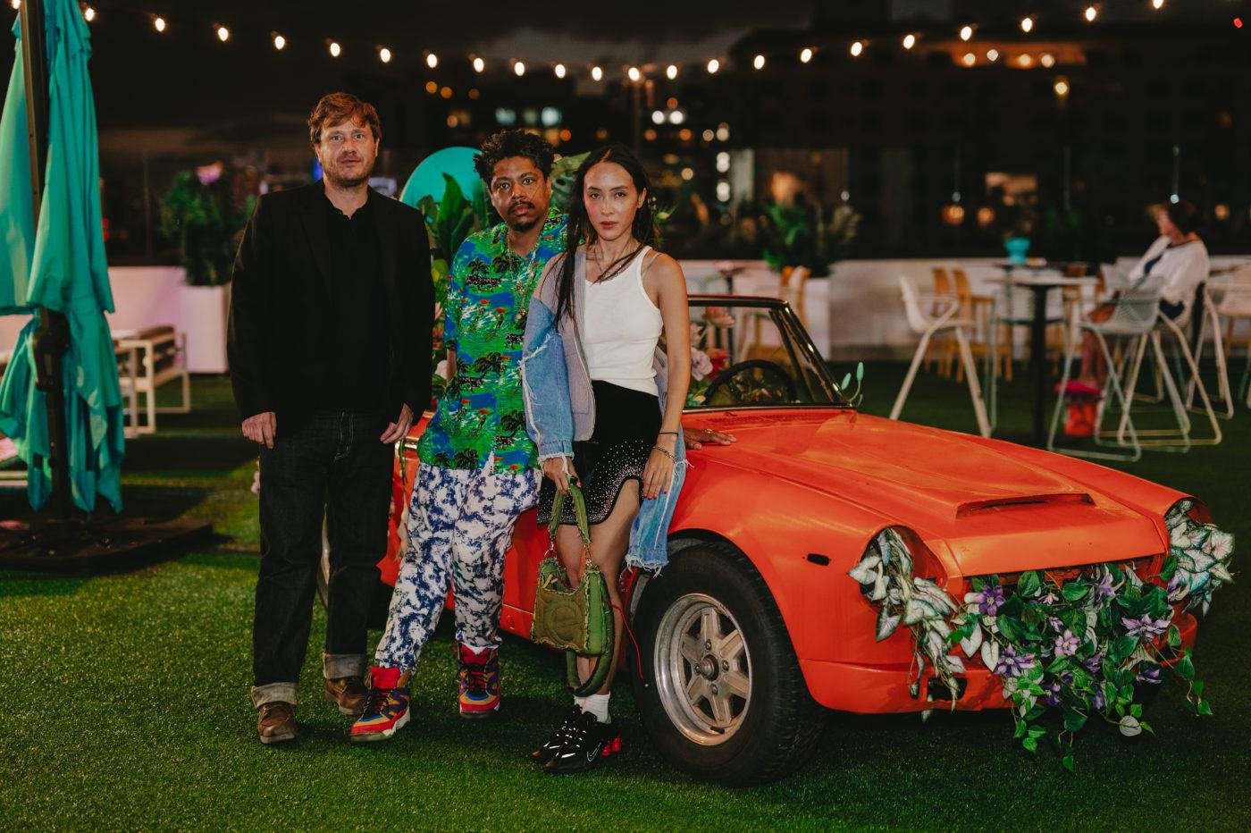 Three people standing near an orange sports car with floral decorations, string lights in the background, outdoor setting.