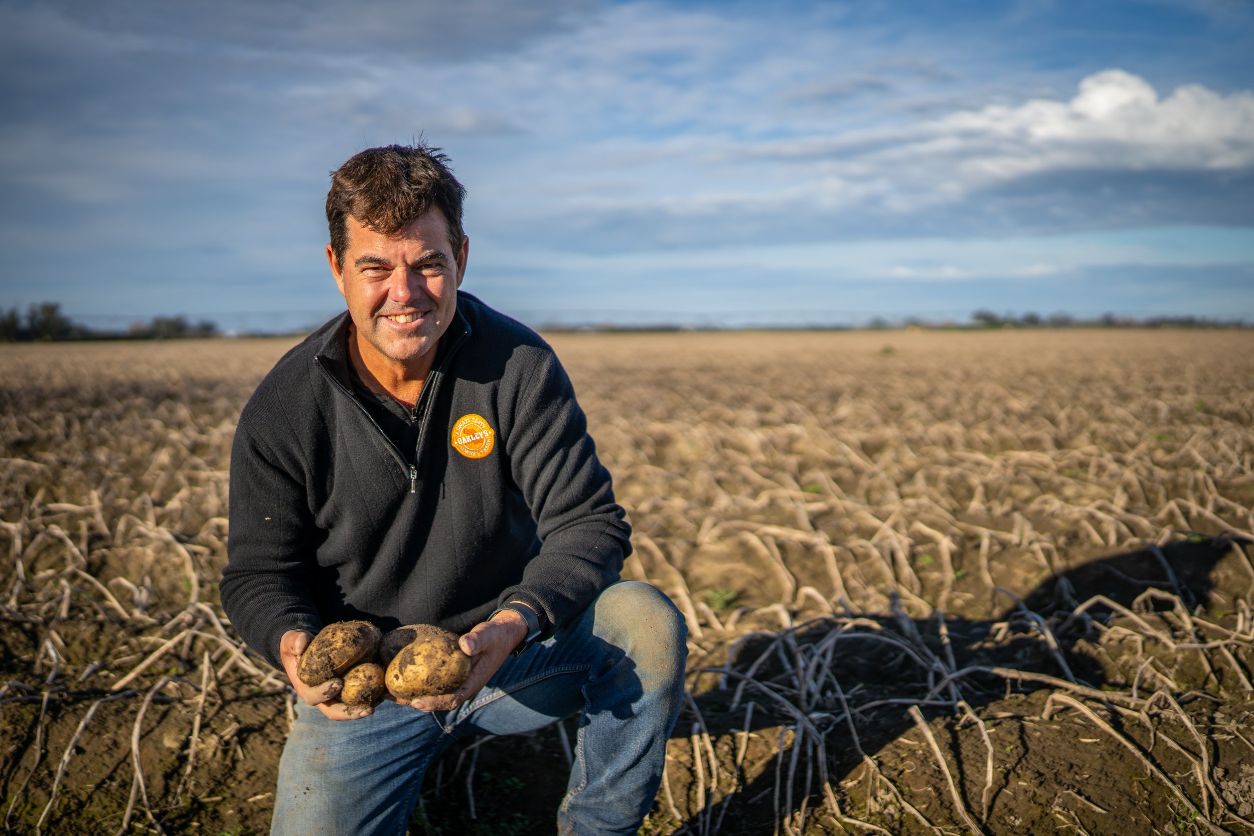 A man kneels in a potato field holding freshly harvested potatoes, with a wide open sky in the background.