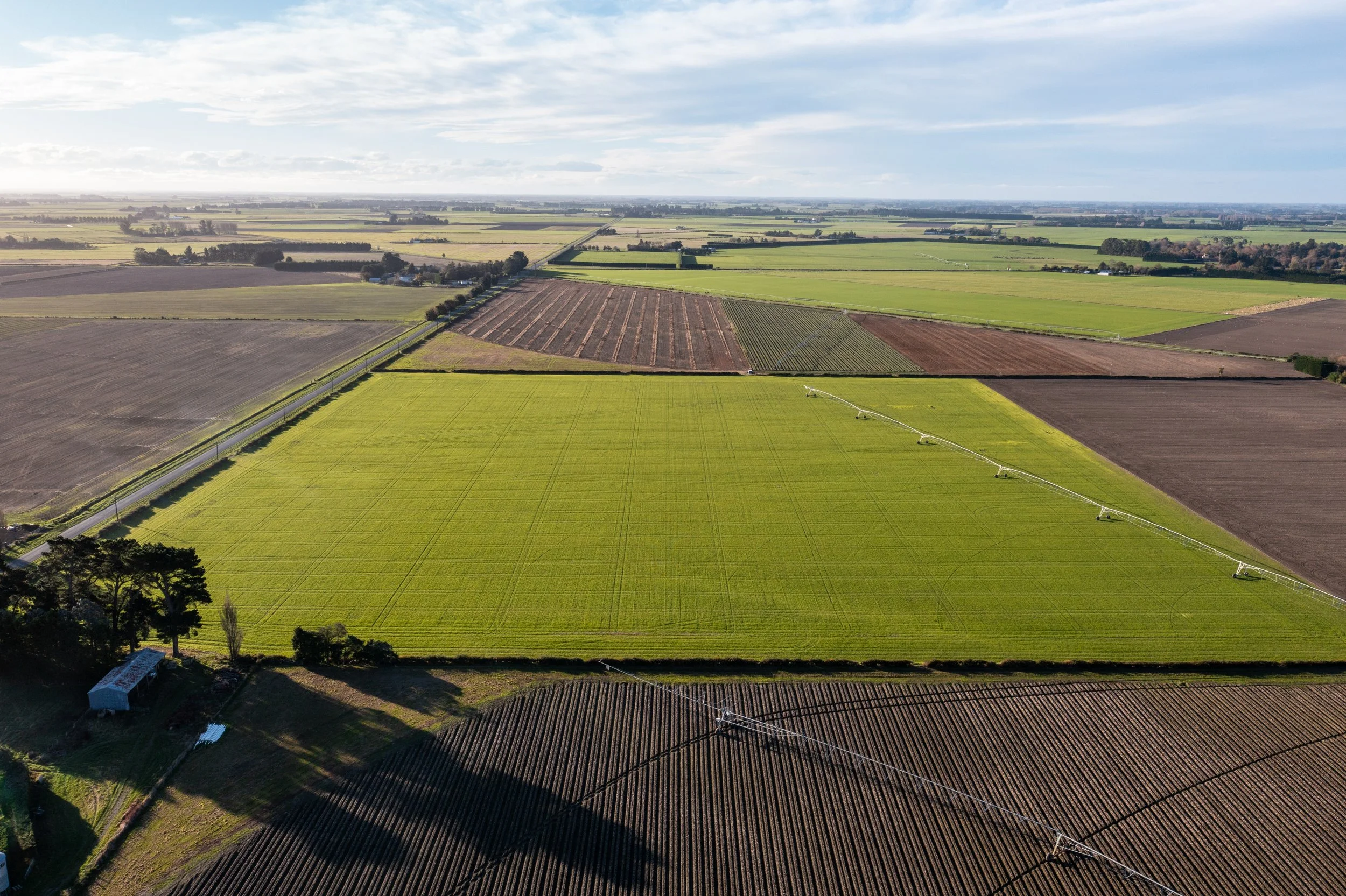 Aerial view of farmland with various fields, farm equipment watering crops, and a small shed at the bottom left, under a partly cloudy sky.
